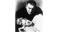 Schwarzweiß-Filmausschnitt: Der in einen schwarzen Umhang gehüllte Christopher Lee beugt sich über eine liegende Frau. Aus seinem Mund fließt Blut. 