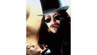 Gary Oldman mit langem Haar, Schnäuzer, roten Augen hinter sehr dunkler Sonnenbrille und mit Zylinder, bildfüllend, hinter/neben ihm verschwommenes Gesicht einer Frau. 