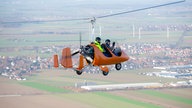 Zwei Menschen fliegen in einem Gyrokopter, im Hintergrund eine Stadt.