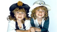 Ashley und Mary-Kate Olsen im Alter von acht Jahren bei einer Fotosession, beide Kinder mit Hut.